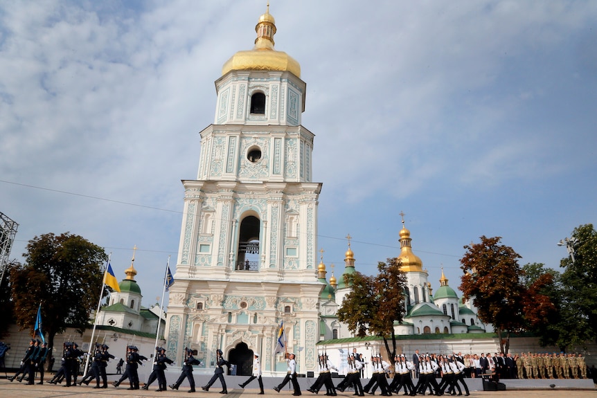 Soldiers in uniform march past a gold topped cathedral.