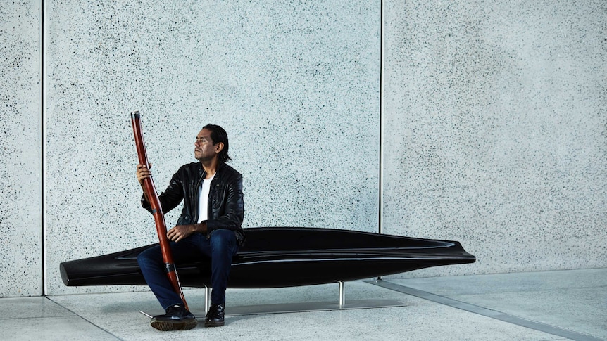 A man sits on the left side of black lounge in front of a concrete wall. He's holding a didgeridoo.