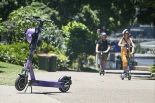 Електронен скутер Neuron, паркиран на пешеходна пътека с двама ездачи на заден план.