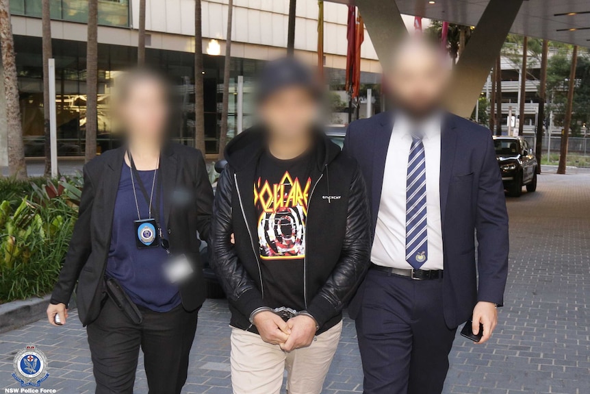 A man with a blurred face being lead through Darling Harbour.