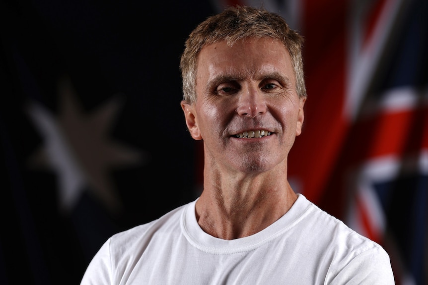 para-triathlete gerrard gosens smiles and poses in front of an australian flag wearing a white t shirt