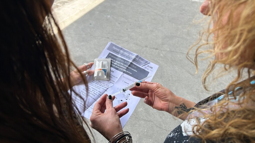 Two women look down at paper instructions and a nasal spray and ampoule of naloxone.