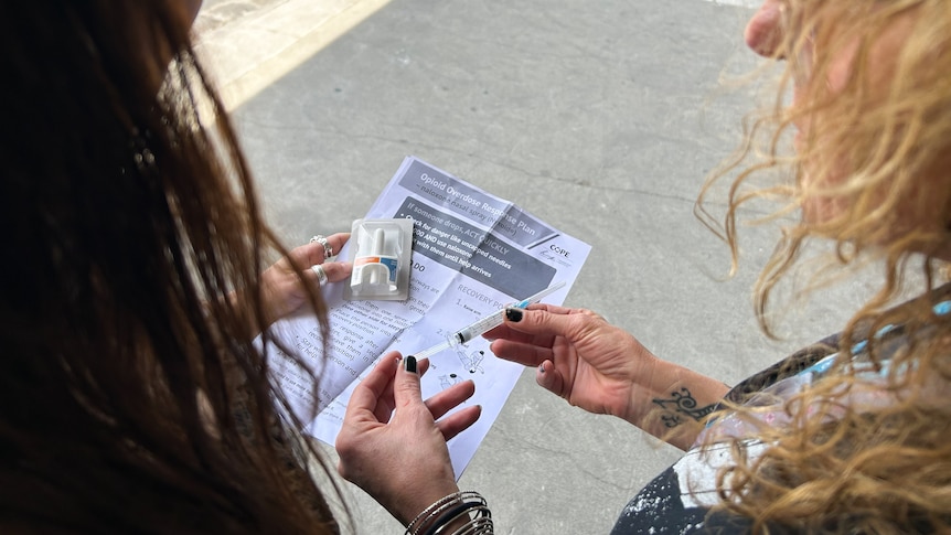 Two women look down at paper instructions and a nasal spray and ampoule of naloxone.