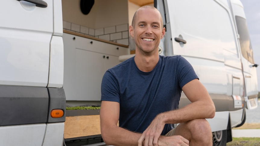 A man crouched down in front of a van's sliding door, he is smiling.
