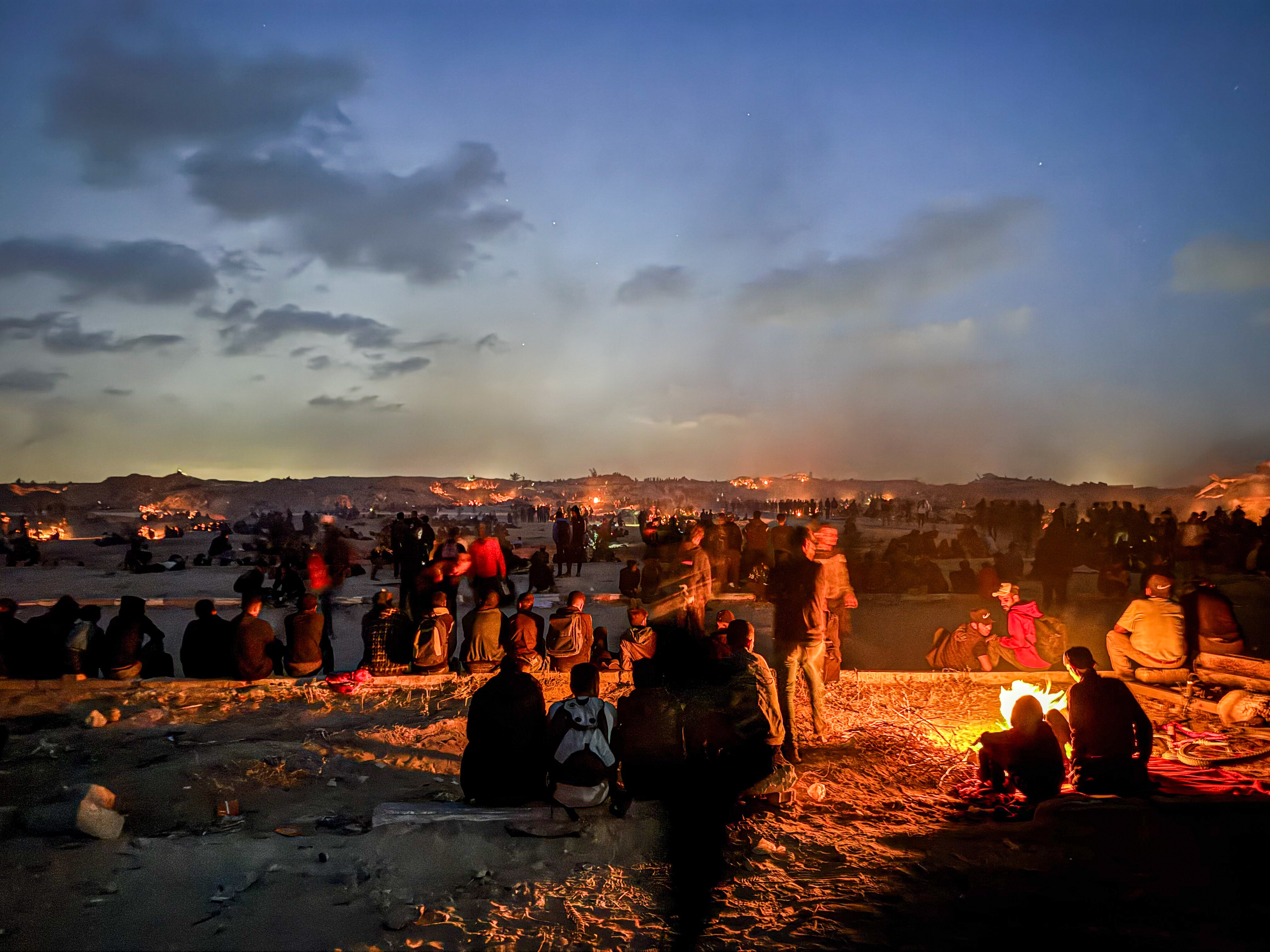 Groups of men sit on the ground around the orange glow of small fires near low hills that stretch into the horizon.