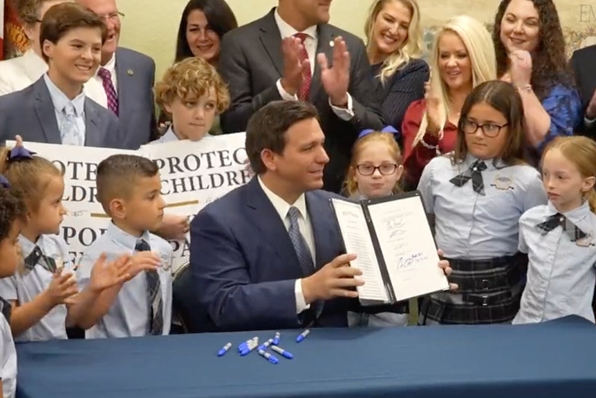 Ron DeSantis sits at a desk and signs papers. Children and adults are gathered behind hi.