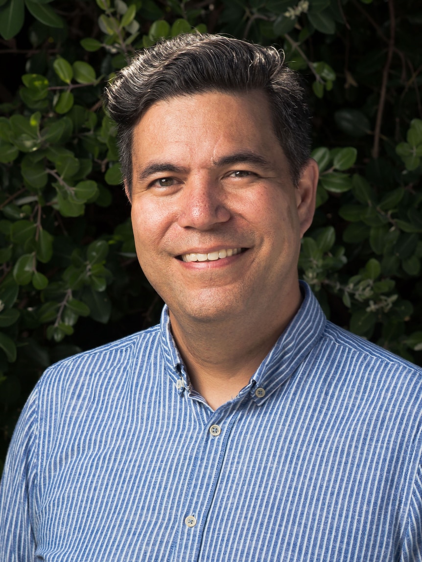 Headshot of a smiling man with brain hair and brown eyes standing in front of green hedge