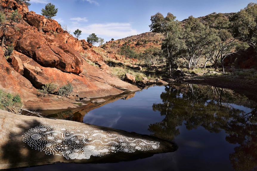A white ochre painting on a flat rock near a waterhole surrounded by red rock and bushland.