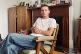 A man wearing a white tshirt and jeans sits with his arms folded on a wooden chair. 