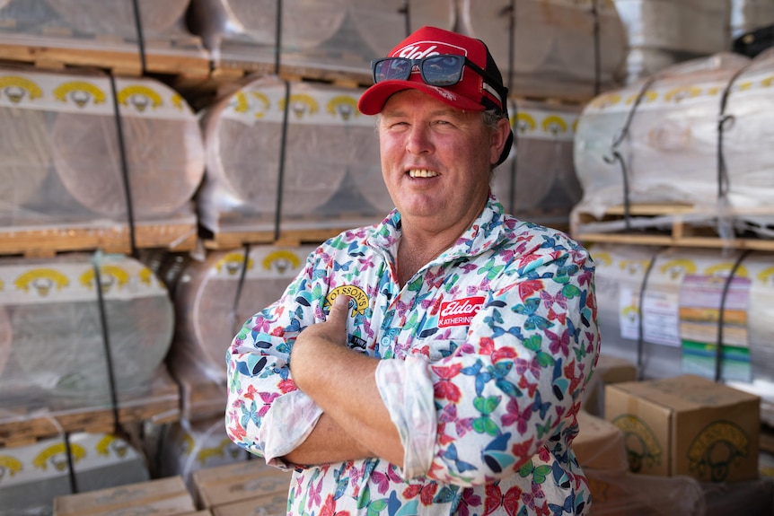 A man wears a red cap and patterned work shirt, looks at the camera with his arms folded, behind him piles of farm supplies.