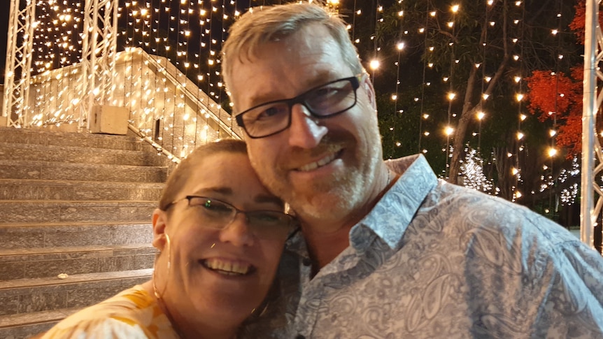 A woman and a man, smiling and hugging with fairy lights in the background.