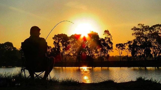 A fisherman silhouetted by the setting sun sits by the river.