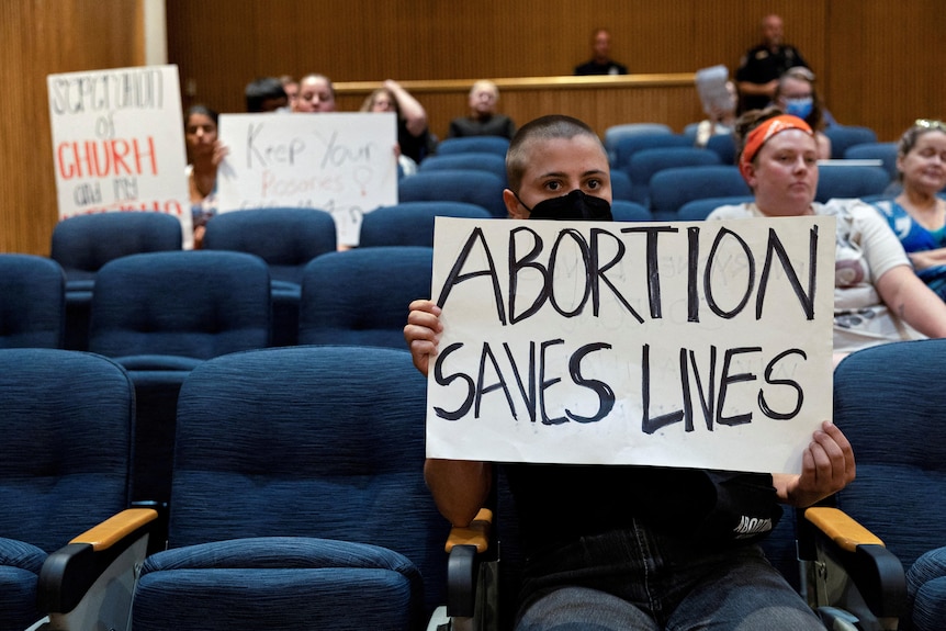 A young woman with a mask and shaved head holds a sign saying "abortion saves lives".