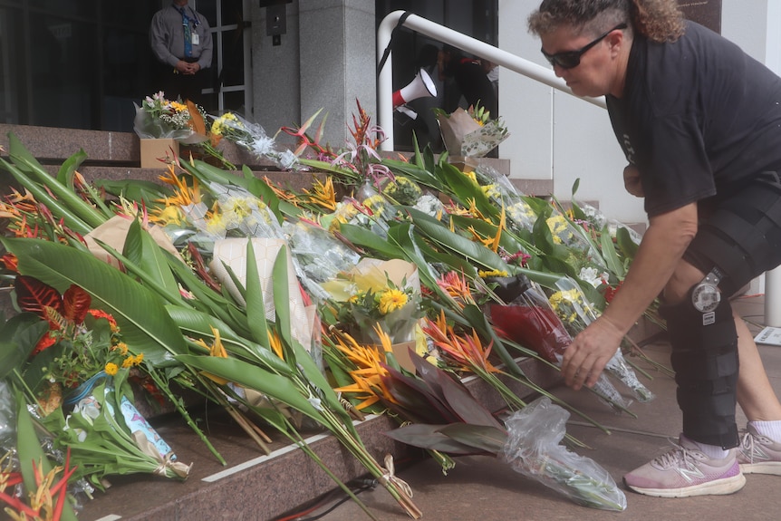 Some flowers left on the steps of a large building, with a person adding another bundle