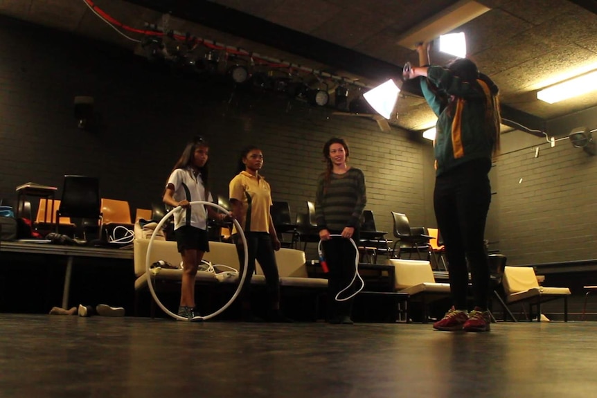 1 girl practicing a performance in a dark school drama room, with 2 other performers and their instructor looking on.