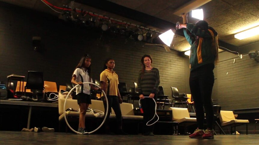 1 girl practicing a performance in a dark school drama room, with 2 other performers and their instructor looking on.