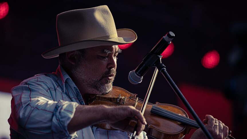 A middle-aged man in a cowboy hat plays violin into a microphone