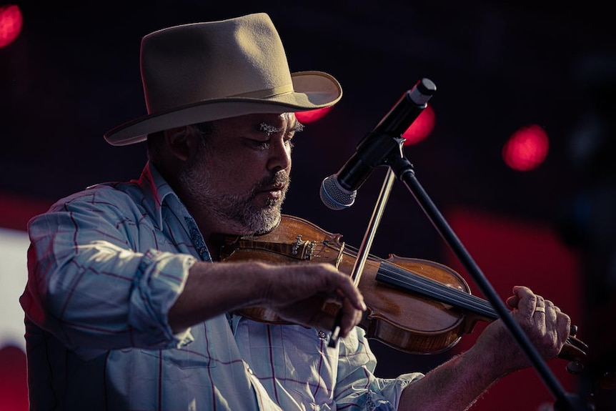 A middle-aged man in a cowboy hat plays violin into a microphone