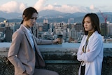 Two women standing and not looking at each other with view of skyline, mountains and clouds in the background. 