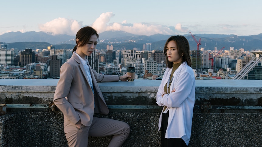 Two women standing and not looking at each other with view of skyline, mountains and clouds in the background. 