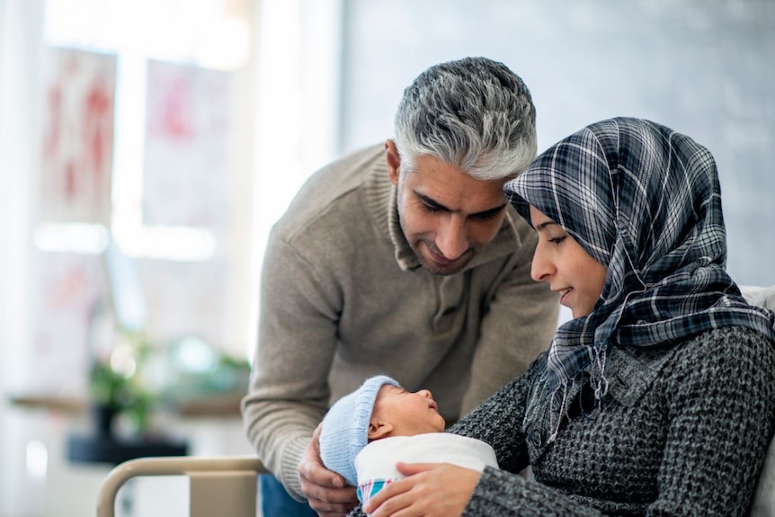 In a light room, a woman cradles a small baby while a man crouches over them. Both adults are smiling.