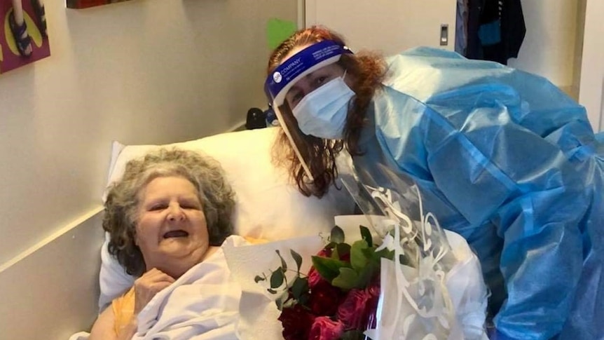 A woman in full PPE smiles for the camera and holds flowers to an older woman in a bed.