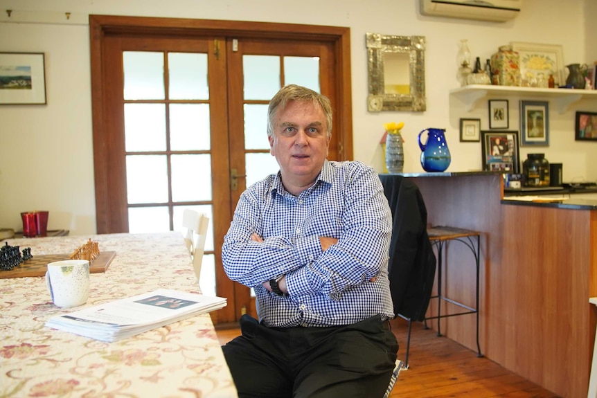 An older man sitting with his arms crossed at a dining table.