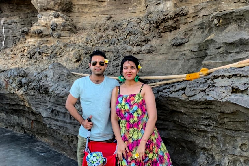 Man and woman standing with pram with small child outside in front of rock face.