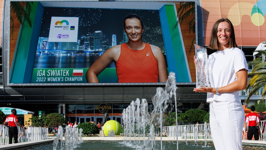 A tennis player poses with the Miami Open trophy - behind her a screen has her name, "2022 Women's champion".