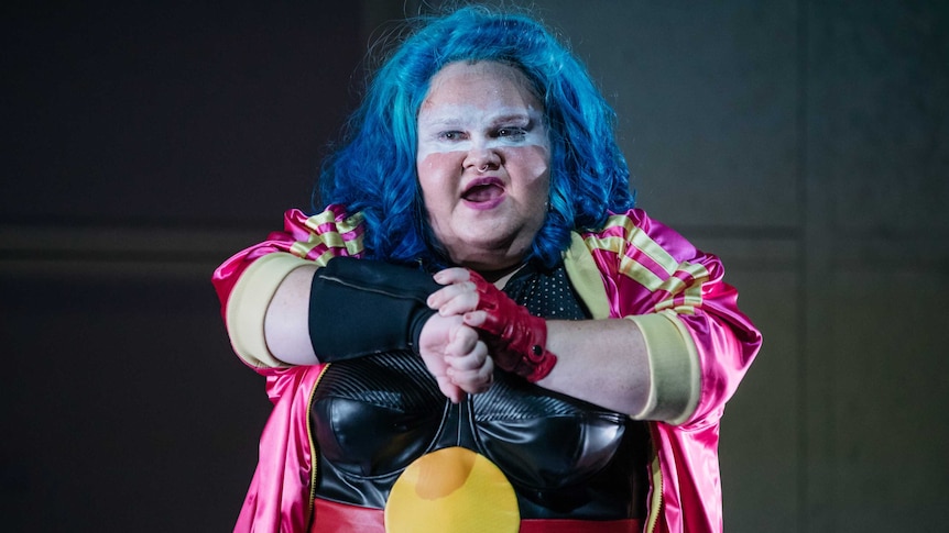 The actor stands, wearing a blue wig, facial makeup and a bustier emblazoned with the Aboriginal flag.