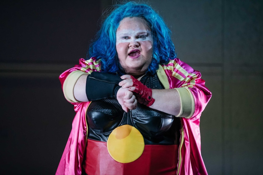 The actor stands, wearing a blue wig, facial makeup and a bustier emblazoned with the Aboriginal flag.
