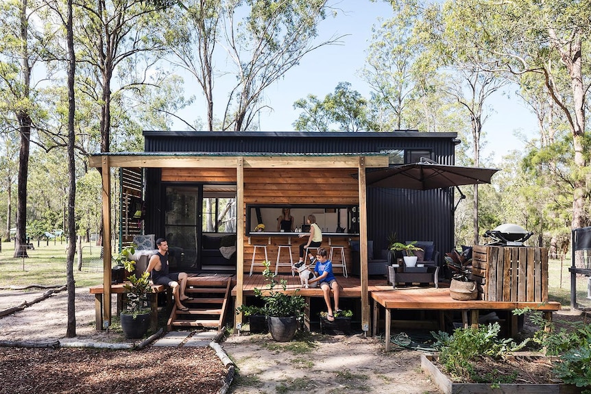 A family of four in front of a black and wood framed small house set amid bush land.   