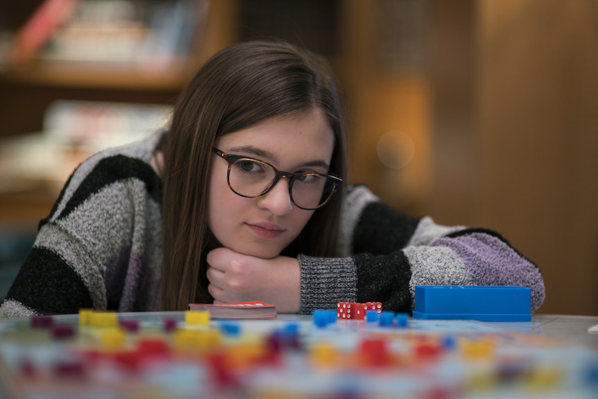 A bespectacled young girl in black and grey striped jumper hunches forward on table near dice and board game pieces.