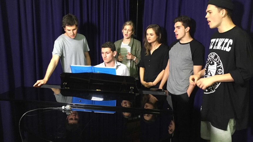 A group of young performers gather around a piano for a song.