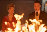 A man and woman in old-fashioned clothing look at something burning in front of them.