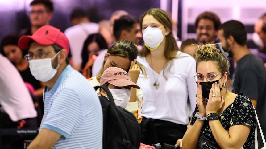 A queue of passengers stand around wearing face masks waiting to board a flight at an airport