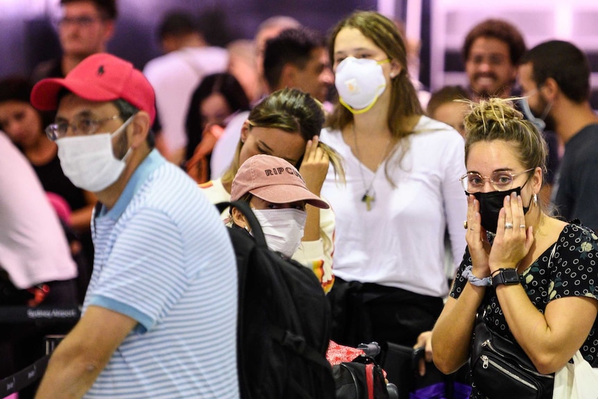 A queue of passengers stand around wearing face masks waiting to board a flight at an airport.