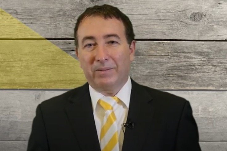 A smiling man in a tie and suit stands in front of a wooden wall