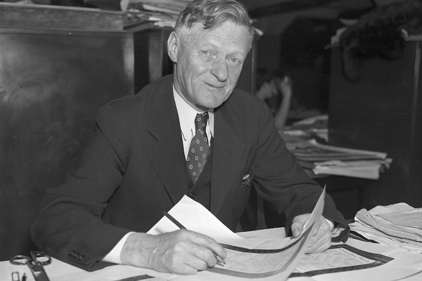Black and white photo of a suited man smiling with mouth closed, sitting at a desk holding large sheets of paper.