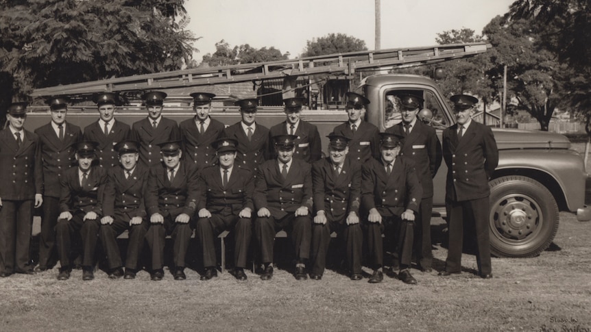 Group of 17 men in rural firefighter dress uniform, posing for a photo in two rows in front of a fire trucker circa 1940s.