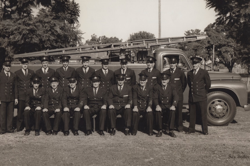 Group of 17 men in rural firefighter dress uniform, posing for a photo in two rows in front of a fire trucker circa 1940s.