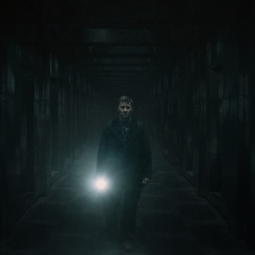 Man in dark clothes is walking in a large dark room and shining a torch straigt ahead at the camera.