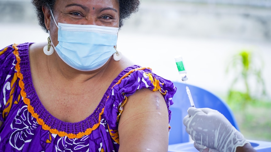 A Papua New Guinean woman in a face mask sits next to a nurse holding a needle