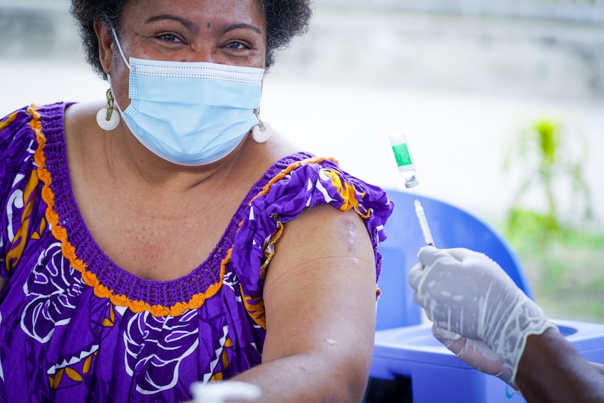 A Papua New Guinean woman in a face mask sits next to a nurse holding a needle