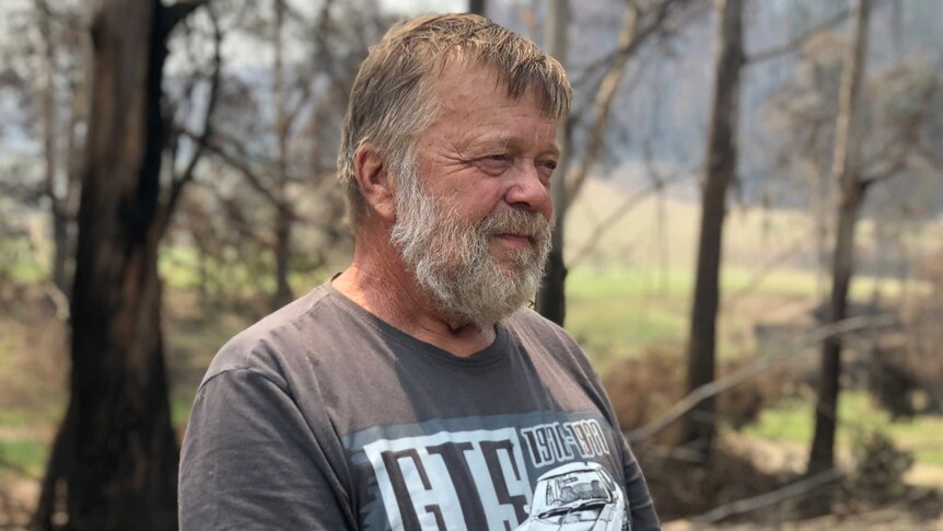 A bearded man standing in front of bush and burnt out trees. He's smiling and not looking at camera