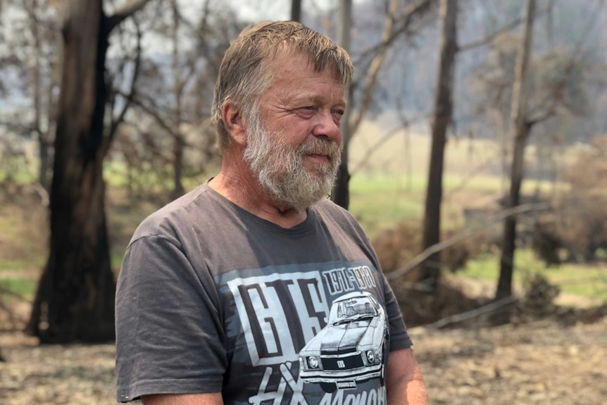 A bearded man standing in front of bush and burnt out trees. He's smiling and not looking at camera