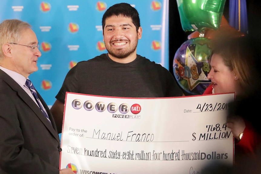 A man smiles as another man in a suit presents him with a giant novelty cheque with his lottery winnings on it.