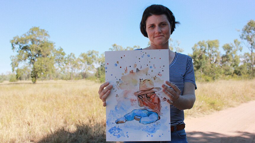 Maree Power stands in the red dirt, holding a watercolour art work of her son.