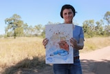 Maree Power stands in the red dirt, holding a watercolour art work of her son.