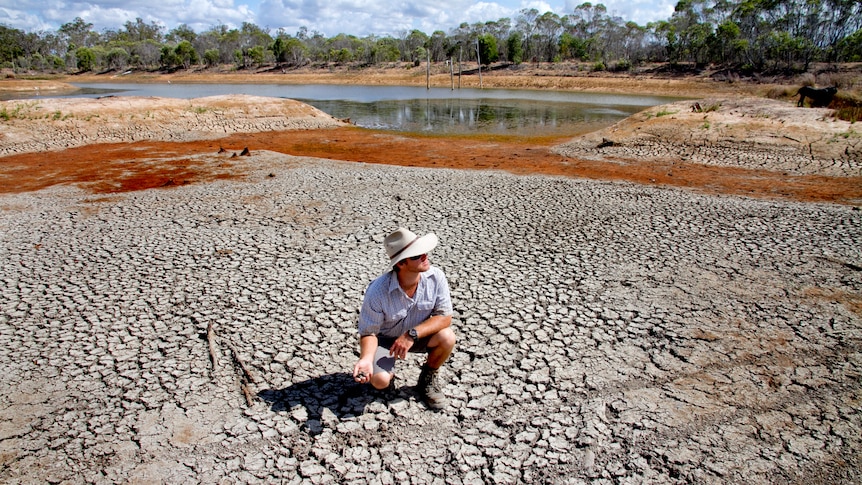 Man in big hat squats in dried up river bed.